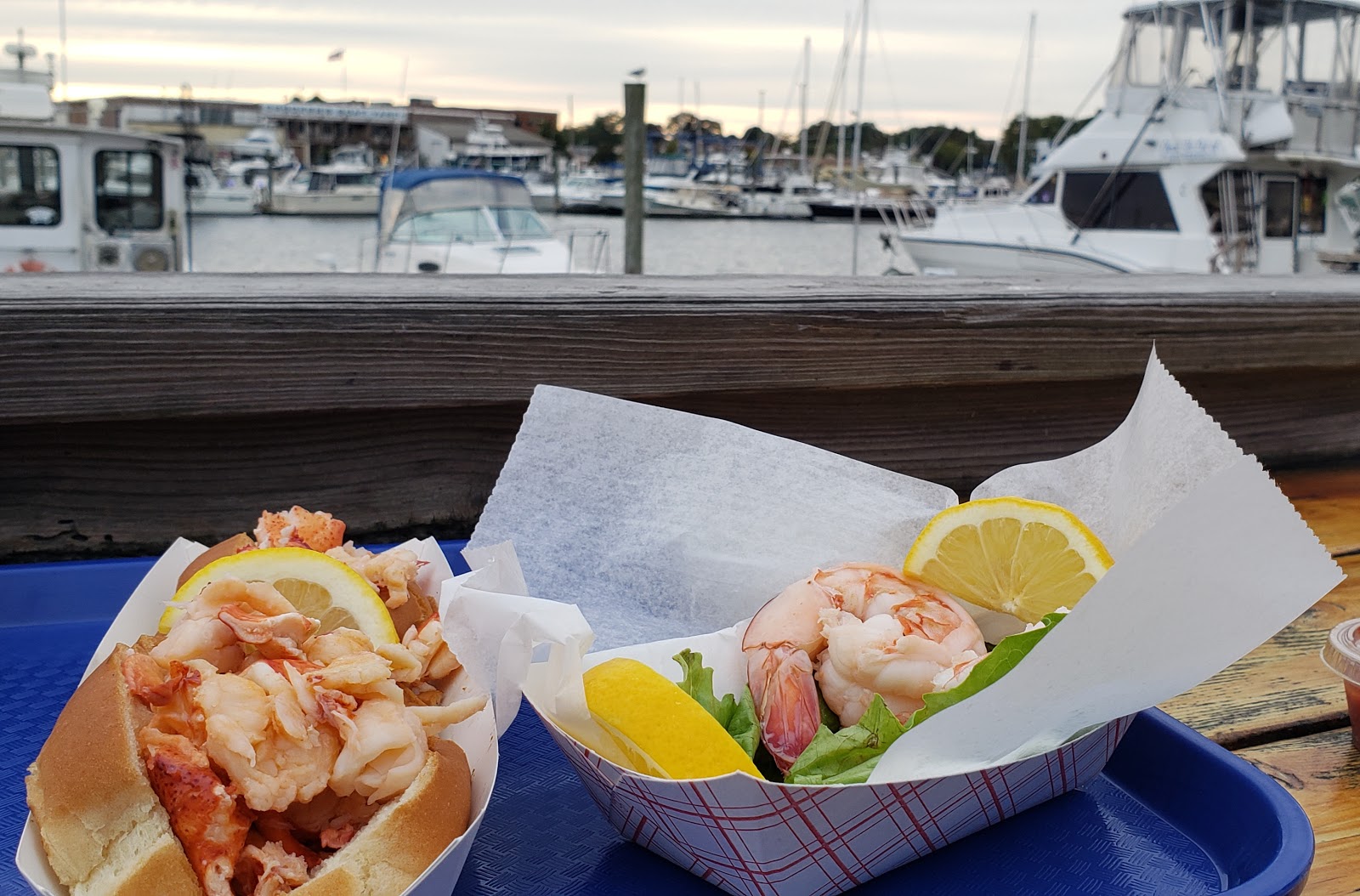 A lobster roll and plate of shrimp sit on a picnic table overlooking boats in the marina