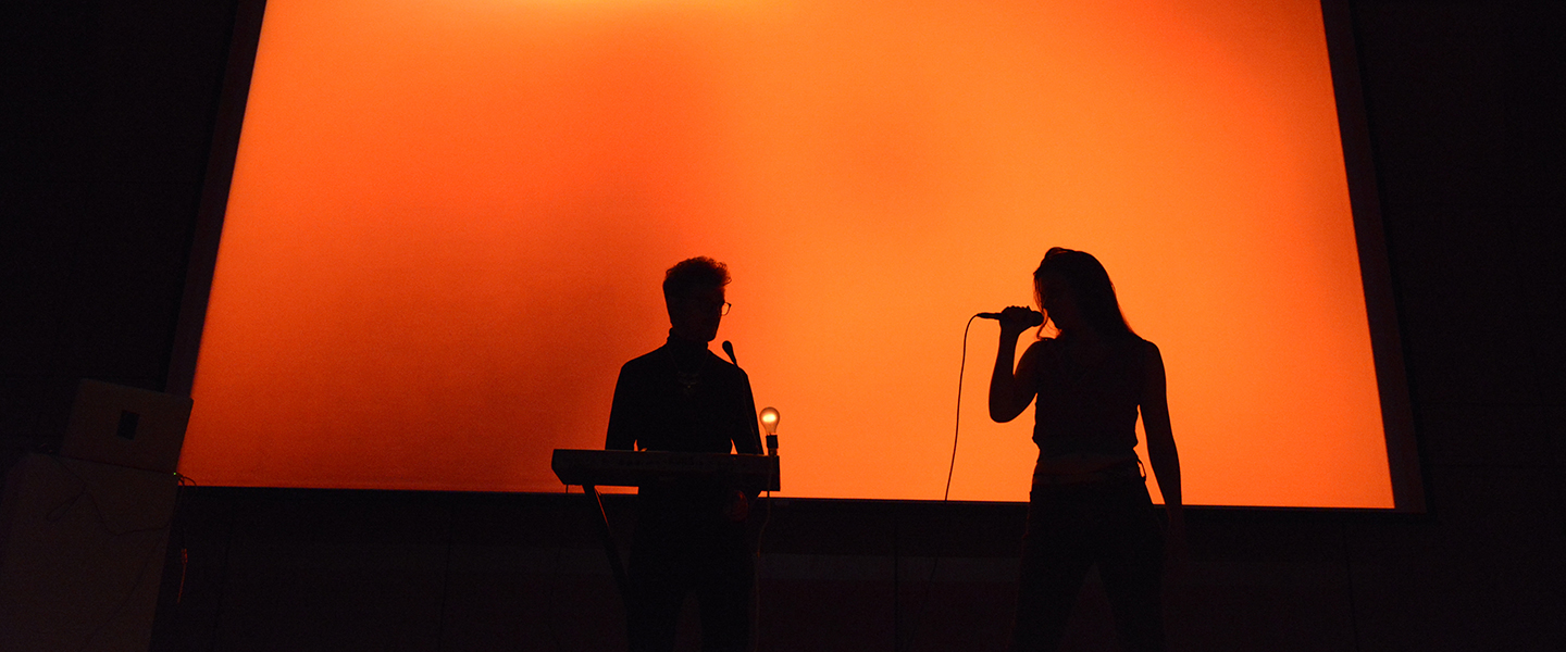 Two singers are silhouetted against a bright orange background
