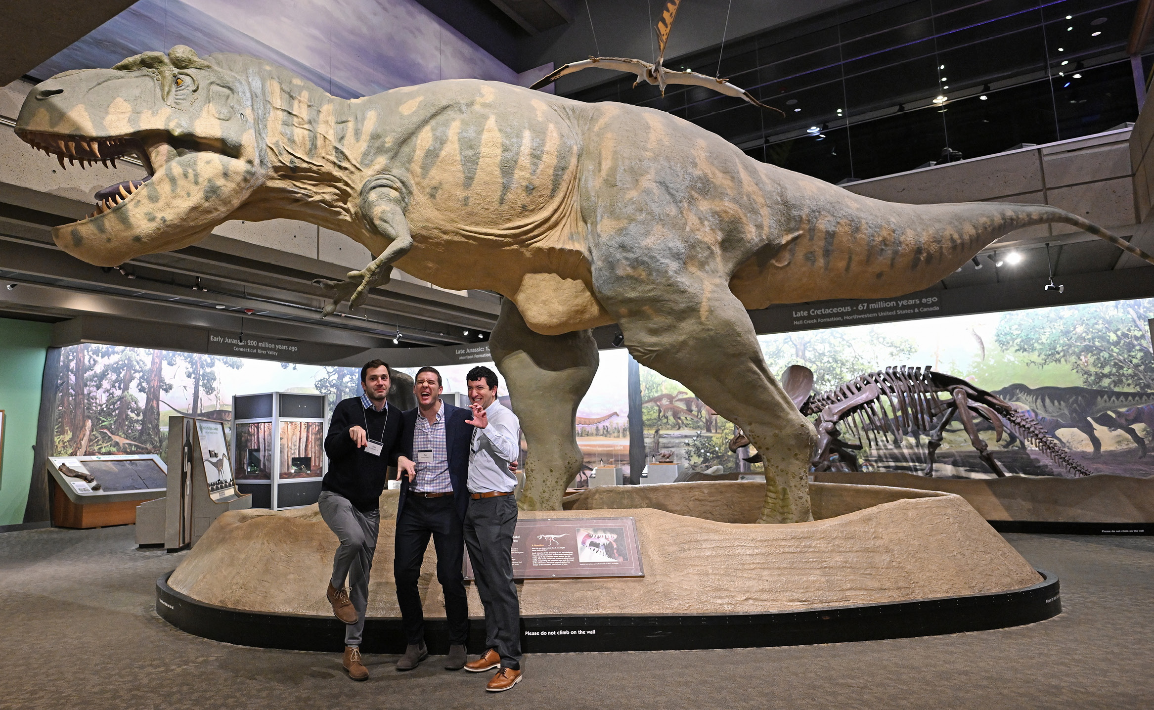 Alums pose in front of a giant dinosaur at an alumni holiday party at the Boston Museum of Science.