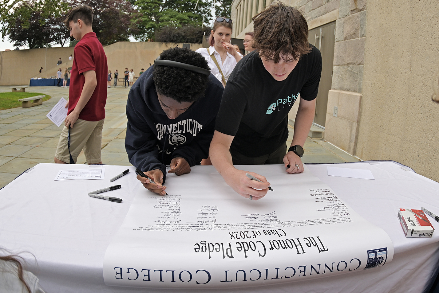 Two students sign a large document at a table in an outdoor courtyard.