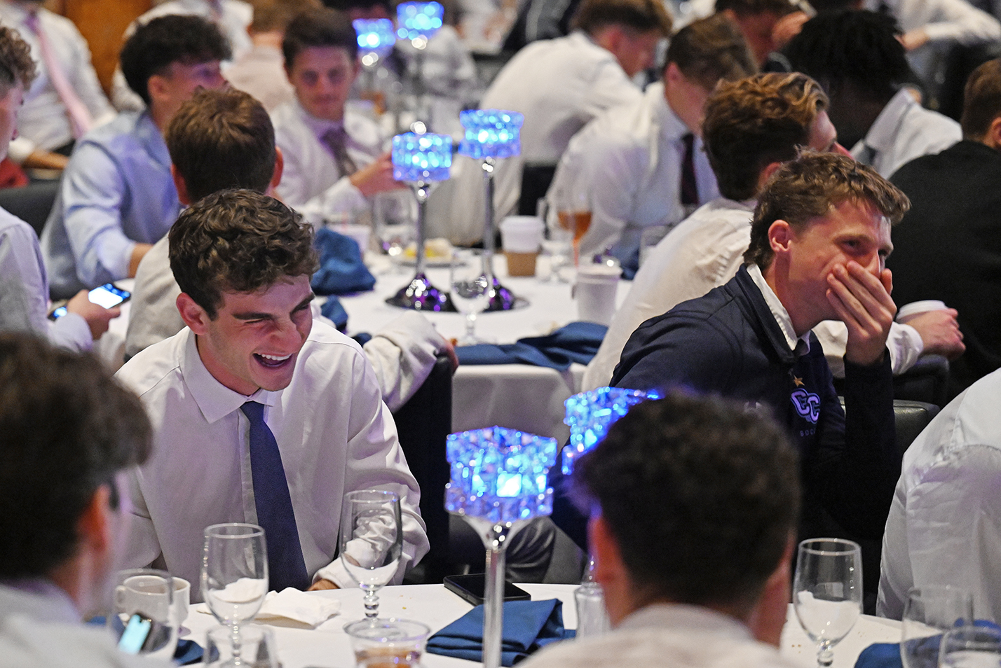 soccer players in dress shirts and ties laugh at a comedian during a team banquet.