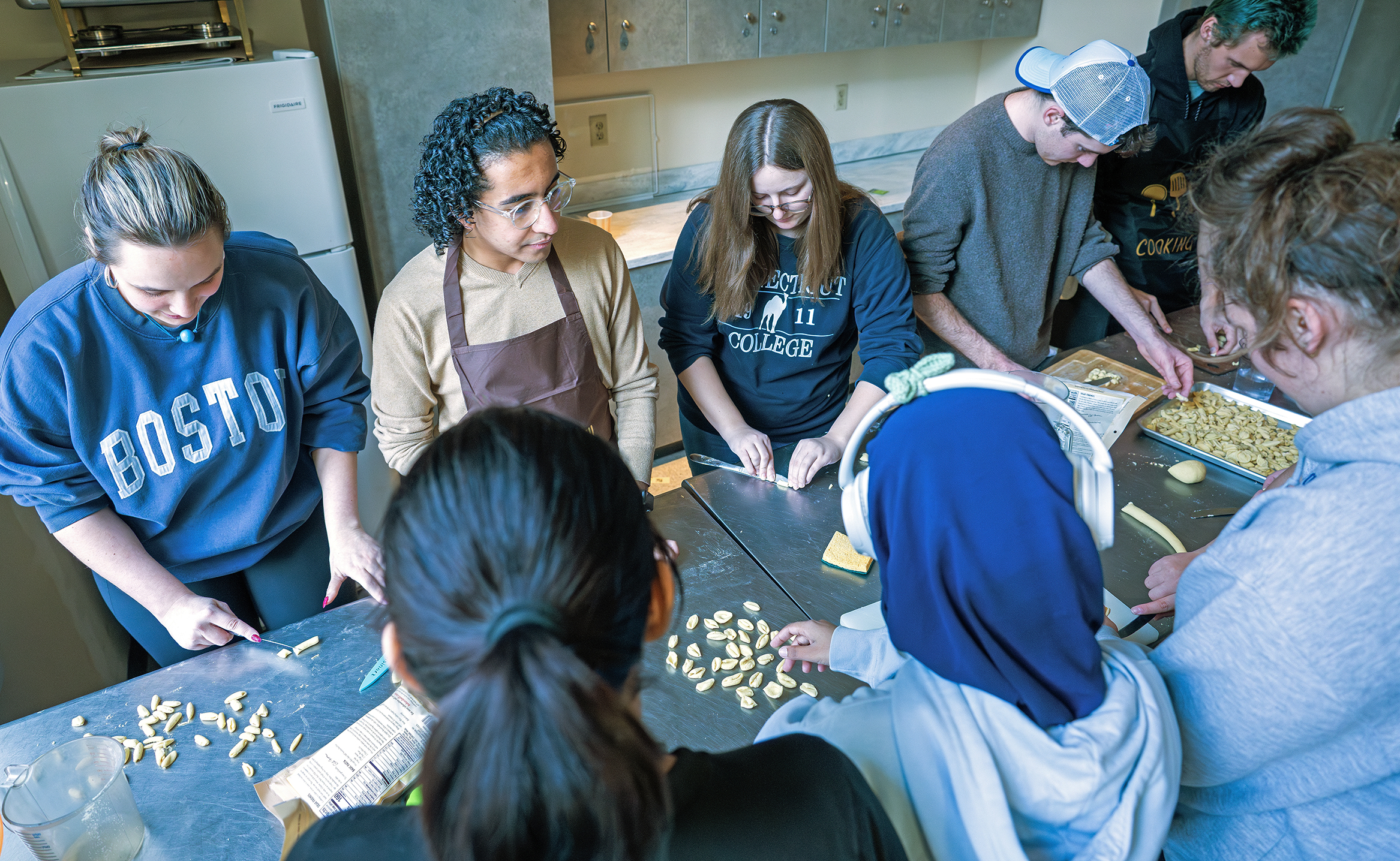 Students in aprons gather around a kitchen work table to make pasta.