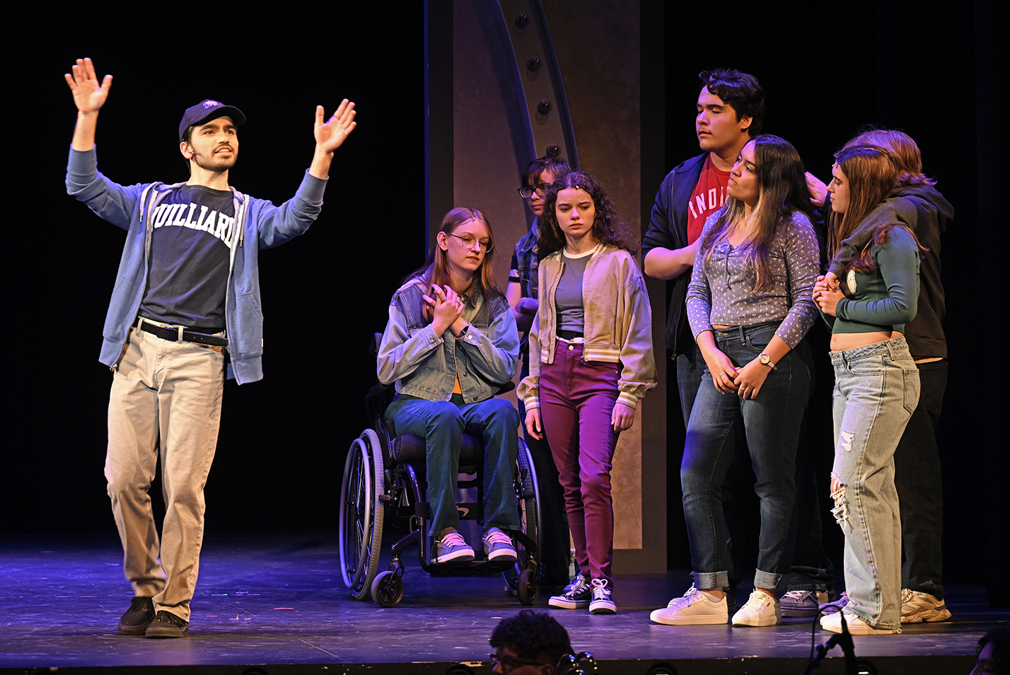 An actor, with arms raised, sings in front of a group of actors on stage during a musical theater performance.