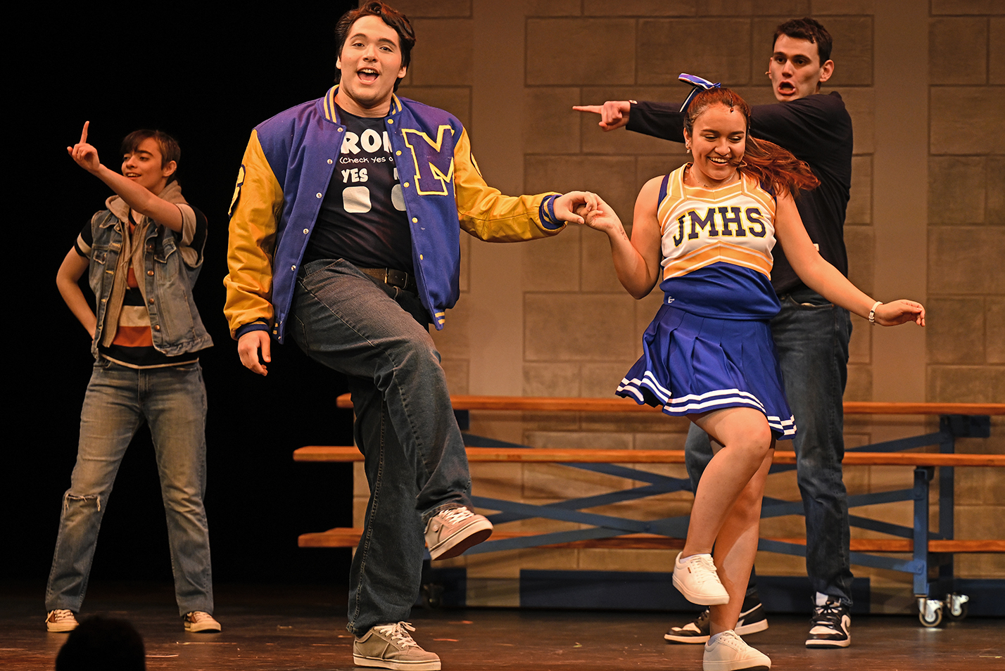 Actors in high school costumes dance on stage during a musical theater performance.